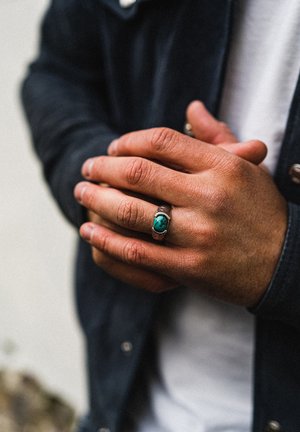 Silver ring with a turquoise stone, worn on a hand with medium skin tone. The hand is partially clasped, showcasing textured skin and jewelry.