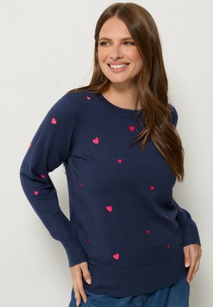 Femme aux longs cheveux bruns souriante, portant un pull bleu marine décoré de petits cœurs roses et un jean bleu.