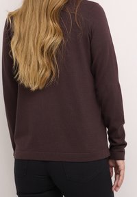 Haut à manches longues couleur bordeaux en tissu texturé, avec un col rond et un ourlet arrondi, associé à un jean noir ajusté.