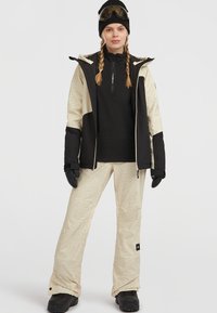 Ski-outfit met een zwarte zip-up top, beige patronen sneeuwbroeken, en een slanke zwarte jas met contrasterende mouwen. Inclusief zwarte handschoenen en een skibril.