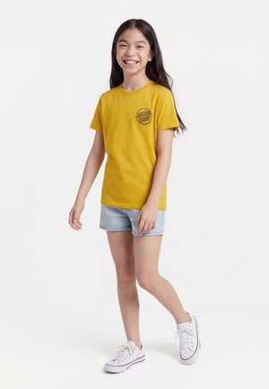 Glunderend meisje met een geel "Santa Cruz" T-shirt, lichte denim shorts en witte sneakers, lopend tegen een effen witte achtergrond.