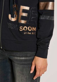 Schwarze Zip-Hoodie mit glänzenden bronzefarbenen Buchstaben und Nieten, die "SEE YOU SOON 5th Ave. NYC." sagen. Glattes Material mit gerippten Bündchen.