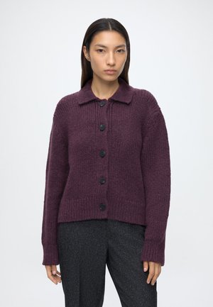 Junge Frau, die eine dunkelviolette, geknöpfte Strickjacke und dunkelgraue Hose trägt, steht vor einem schlichten hellen Hintergrund.