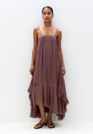 Maxi dress in soft, mauve fabric with thin straps, a flowing silhouette, and uneven hemline featuring frayed edges. Worn with flat sandals.