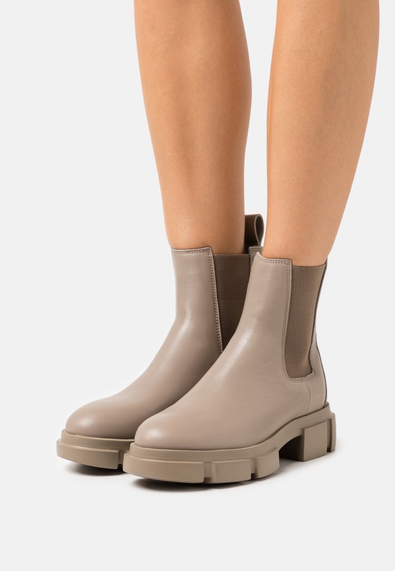 Bottines en cuir taupe avec semelles épaisses et panneaux latéraux élastiques, portées sur des jambes nues sur un fond clair uni.