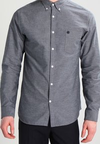 Pánská košile na knoflíky v tmavě modro-bílém psím zubu, vybavená náprsní kapsou s logem a klasickým límcem.
