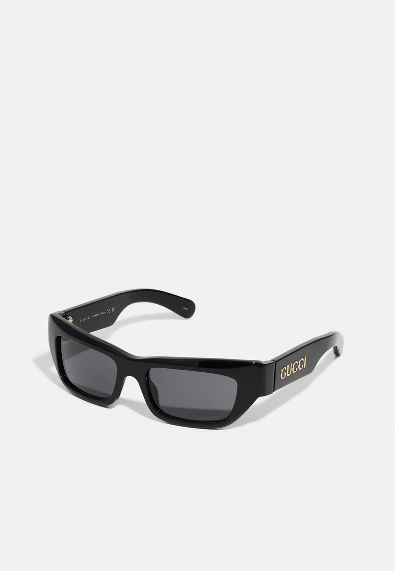 Black sunglasses with a thick acetate frame, gray lenses, and gold "GUCCI" logo on the sides. Features a square shape and smooth texture.