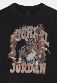 Juodas marškinėliai su trimis krepšininkais veiksme, pavaizduotais grafikoje. "MICHAEL JORDAN" tekstas ryškiai išsiskiria raštu, primenančiu senovines reklamines formas.