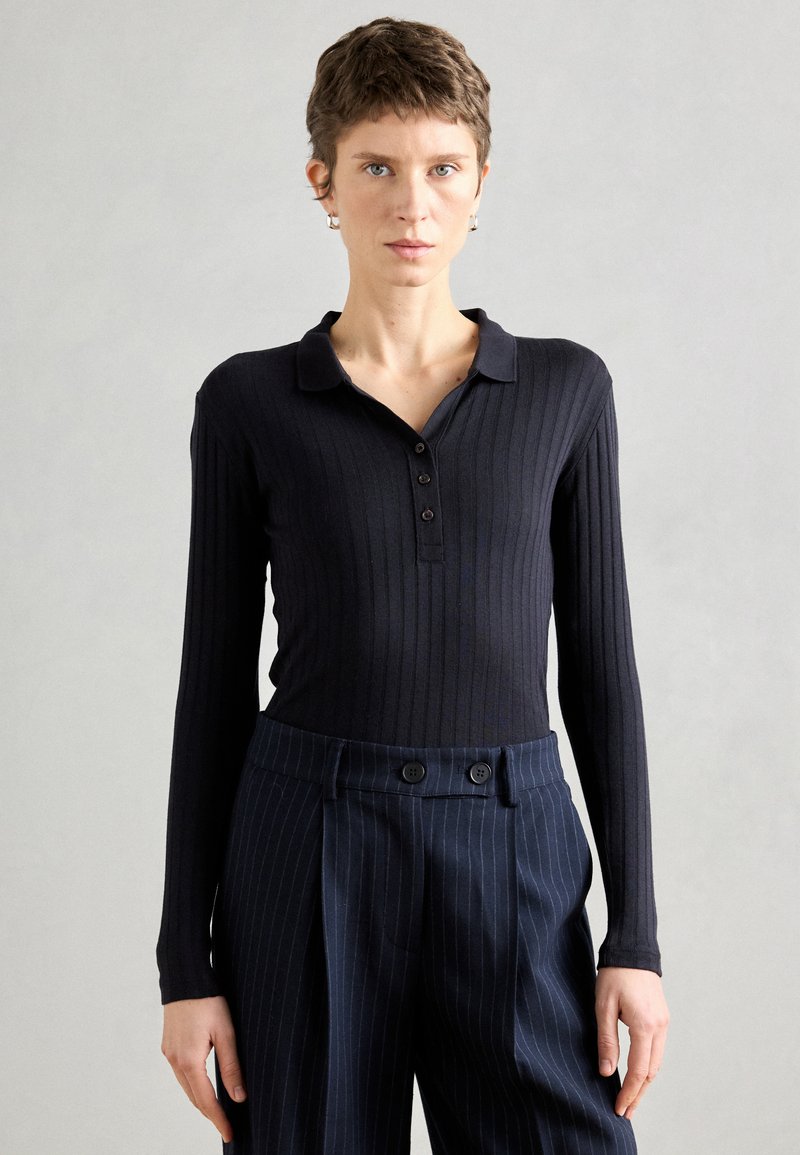 Top de manga longa riscado preto com gola e fecho de botão, combinado com calças azul marinho com riscas e botões duplos.