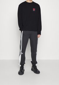 Männlicher Model trägt ein schwarzes Langarm-Hugo-Logo-Shirt, schwarze und graue Trainingshose mit weißen Streifen und schwarze Sneaker, steht vor einer weißen Wand.