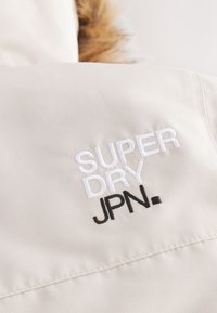 Light beige fabric with soft texture features white and black embroidered text: "SUPER DRY JPN." Accents include a fur trim.