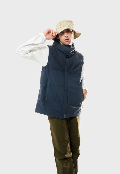Marinblå pufferväst med hög krage, frontdragkedja och sidofickor, bärs över en vit långärmad skjorta och olivgröna byxor. Kaki bucket hat.