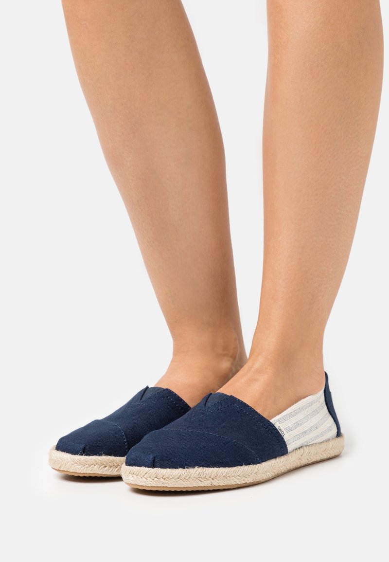 Jambes portant des espadrilles en toile bleu marine à enfiler avec des semelles en corde beige et un tissu rayé blanc à l'arrière sur fond blanc.