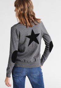 Grau gestrickter Pullover mit einem auffälligen schwarzen Stern auf dem Rücken und kontrastierenden schwarzen Ellbogenpatches. Schlichtes, tailliertes Design mit rippenförmigen Säumen.
