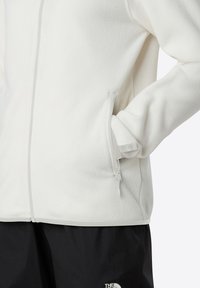 Jachetă albă din fleece cu o textură netedă, buzunare cu fermoar gri și margini gri contrastante pe cusături, asortată cu pantaloni negri.