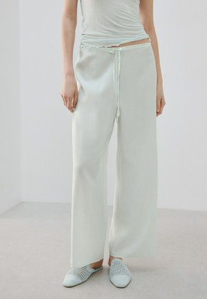 Persona vistiendo pantalones sueltos y claros con cordón, zapatos tejidos tipo slip-on a juego y una camiseta blanca corta en un espacio interior minimalista.