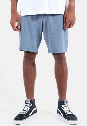 Homme portant un short décontracté bleu clair, un T-shirt blanc et des baskets montantes noires à lacets blancs, debout devant un fond blanc.