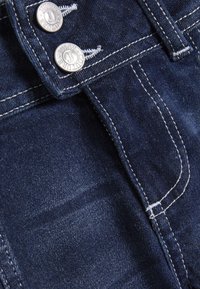 Σκούρο μπλε ύφασμα τζιν με εμφανείς ραφές, δύο ασημένια κουμπιά στη μέση και διακριτικές υφές.