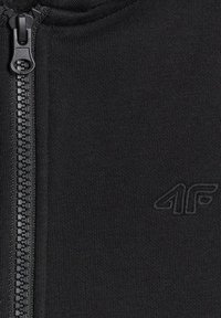 Gros plan sur un tissu noir avec une fermeture éclair noire à gauche et un logo "4F" subtilement brodé sur le côté droit.