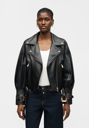 Kvinde iført en sort oversized læderbikerjakke med guldlynlåse og spænder, hvid top og mørkeblå jeans, stående mod en ensfarvet baggrund.