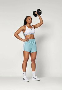 Sutiã desportivo branco com logotipo, calções azuis claros com listas laterais e sapatilhas brancas. A segurar um haltere preto acima da cabeça.