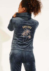 Tuta in velluto blu scuro con colletto alto, caratterizzata da ricami "CAYO COCO" e palme sulla schiena. Tessuto liscio, design aderente.