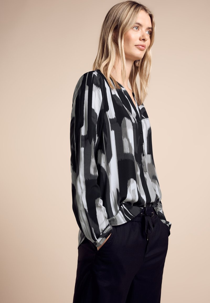 Blouse à motifs noir et gris avec une coupe ample, décolleté boutonné et manches longues, associée à un pantalon noir. Texture de tissu lisse.