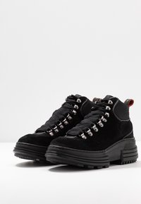Botas de ante negro con suelas de goma gruesas, que presentan una lengüeta de malla texturizada, ojales metálicos para los cordones y una lengüeta naranja en el cuello.