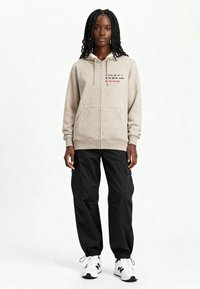 Haut à capuche beige avec fermeture éclair et texte brodé, associé à un pantalon cargo noir et des chaussures de sport blanches. Présente un tissu doux et texturé.