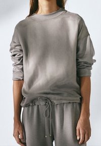 Grijze trui met ronde hals, verzakte schouders, elastische zoom en een trekkoord. Zachte stof met een gladde textuur en een subtiel verloop patroon.