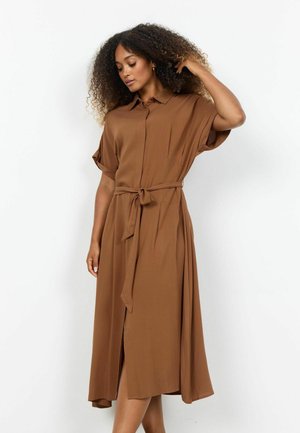 Kvinde med krøllet hår iført en brun kortærmet midikjole med et knyttet bælte i taljen, stående foran en hvid baggrund.