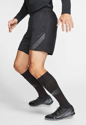 Mann trägt schwarze Nike-Sportshorts, lange schwarze Socken und schwarze Nike Hallenfußballschuhe in einer athletischen Haltung.