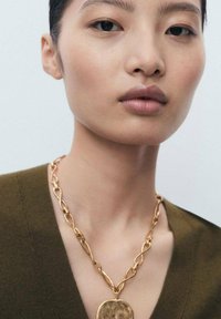 Gold chain necklace featuring elongated, twisted links and a round, textured pendant, worn over a deep olive green V-neck top.
