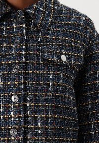 Giacca tessuta di colore blu scuro, con macchie multicolori. Tessuto testurizzato con cuciture visibili e bottoni argentati lucidi.