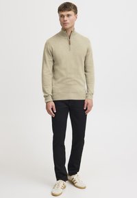 Suéter beige con cuello zip y dobladillo acanalado, combinado con pantalones negros ajustados y zapatillas blancas con detalles en marrón.