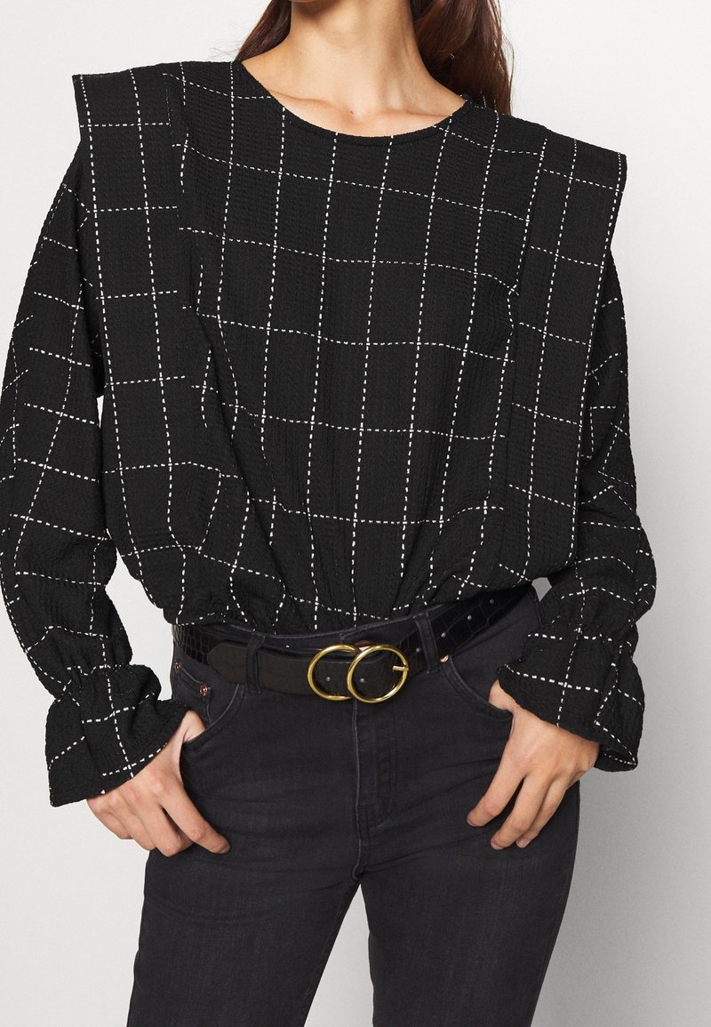 Svart långärmad topp med vitt rutmönster, texturerat tyg och puffärmar, i kombination med högmidjade svarta jeans och ett bälte med dubbel cirkel.