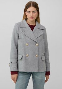 Manteau gris croisé avec de grands revers, boutons dorés et poches avant. Le tissu a une finition texturée. Stylé avec un pull bordeaux.