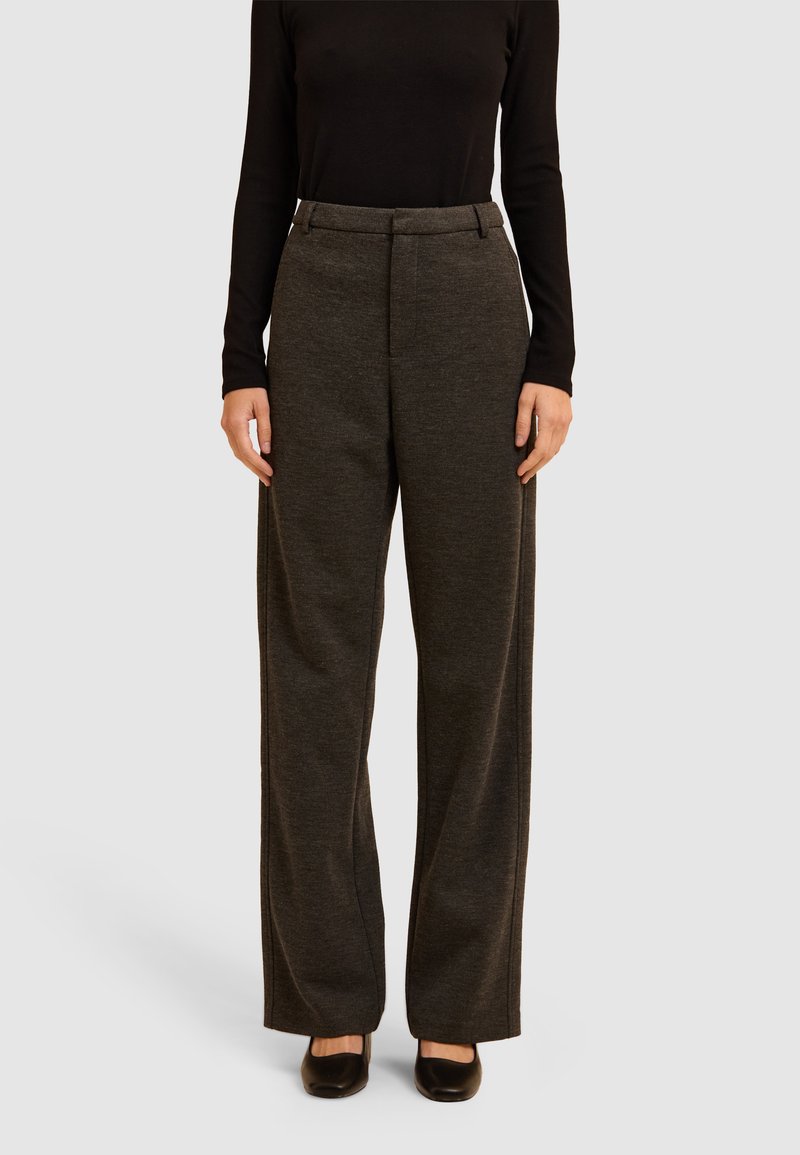 Pantalons larges gris foncé en tissu texturé, avec une taille ajustée et des pinces subtiles à l'avant, associés à des chaussures noires.
