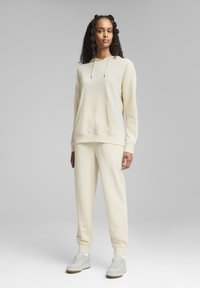 Crème-kleurige hoodie en bijpassende joggers gemaakt van zachte stof. De hoodie heeft een voorzak en een tekstversiering. Sneakers zijn wit met tan-kleurige zolen.