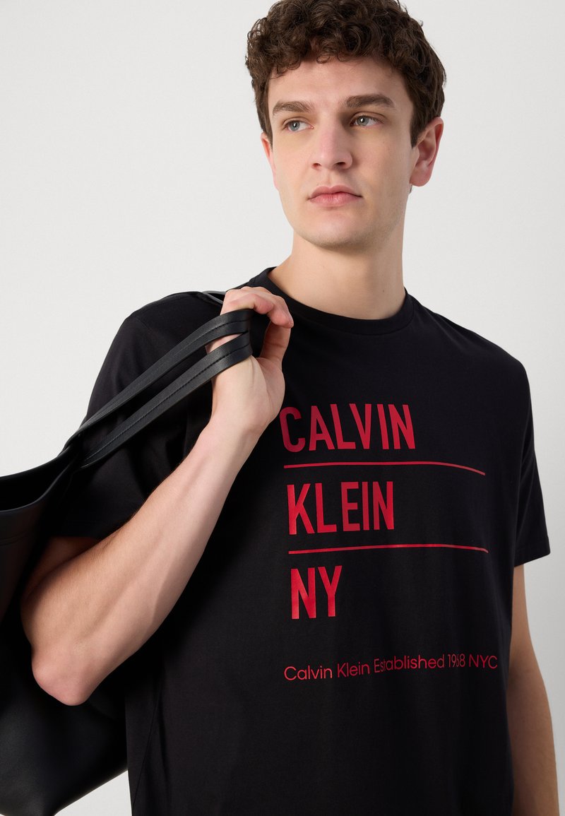 Camiseta de algodón negra con texto impreso en rojo "CALVIN KLEIN NY" y "Calvin Klein Established 1968 NYC", llevada con un bolso negro.