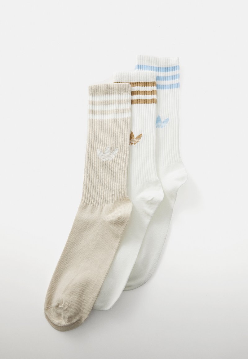 Três pares de meias caneladas: beje com riscas castanhas, brancas com riscas azul claro e brancas sólidas. Cada uma tem um pequeno detalhe de logótipo.