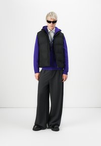 Czarna kamizelka puchowa na purpurowym hoodie, szare szerokie spodnie i czarne buty na platformie. Modelka nosi okulary przeciwsłoneczne. Minimalistyczne tło.