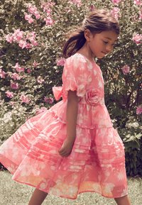 Roza cvetlična obleka s kratkimi napihnjenimi rokavi in plastmi volanov. Na pasu je poudarek v obliki vrtnice. Prosojen, lahek material z živahnim vzorcem.