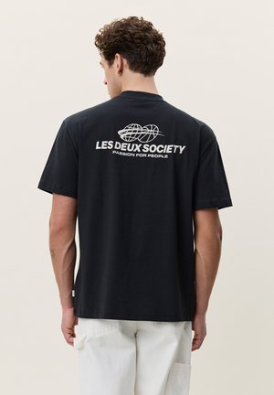 Homme aux cheveux bouclés portant un T-shirt noir avec le texte blanc « LES DEUX SOCIETY PASSION FOR PEOPLE » et des graphismes, assorti à un pantalon blanc.