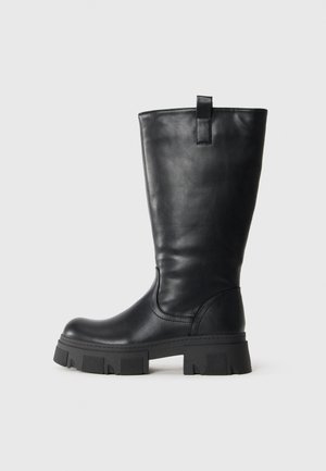 Bota de cuero negra hasta la mitad de la pantorrilla con suela gruesa de tacos y lengüeta en el costado, mostrada de perfil sobre fondo blanco.