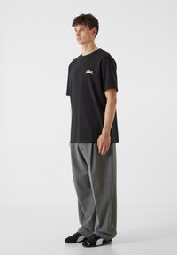 Melna kokvilnas T-krekls ar brīvu piegriezumu, ar mazu zelta logo kreisajā krūtī; kopā ar vaļīgiem pelēkiem biksēm un melniem kedām.