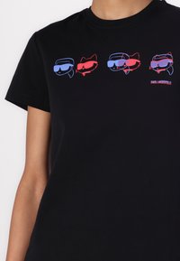 Black t-shirt with crew neck, featuring three cartoon-like figures in blue and red glasses. "Karl Lagerfeld" logo in red on the bottom right.