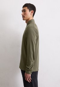 Polo a maniche lunghe verde oliva con colletto strutturato, polsini con bottoni e orlo dritto. Il modello lo indossa con jeans scuri.
