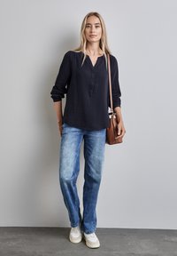 Camicia button-up blu navy con maniche lunghe, tessuto testurizzato; abbinata a jeans azzurri chiari e sneakers bianche; borsa a tracolla marrone.