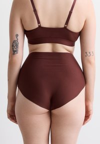 Person, der braune hochtaillierte Unterwäsche und einen passenden BH trägt, mit Tattoos an den Oberarmen, steht vor einem schlichten Hintergrund.